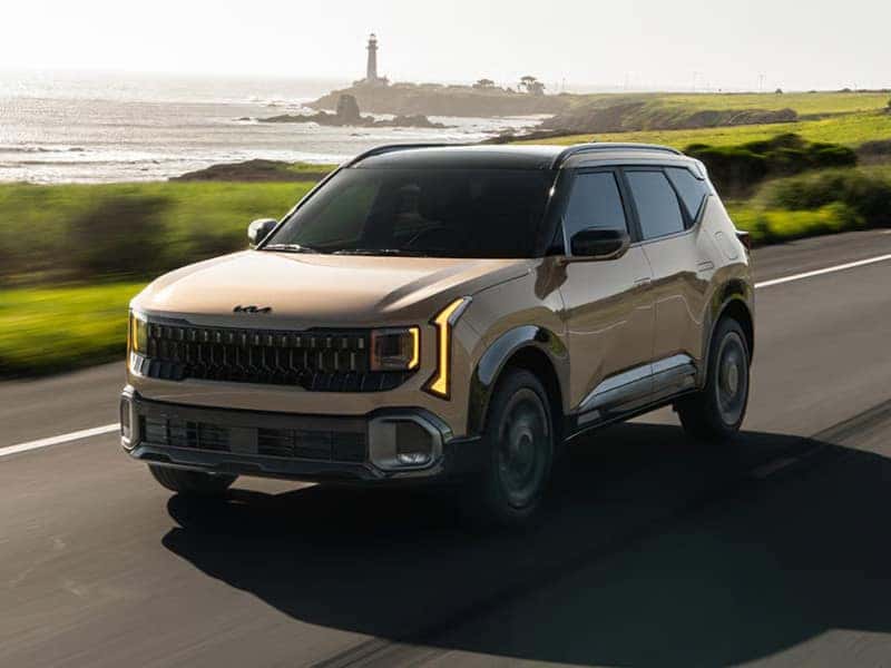 A tan SUV drives along a coastal highway with a lighthouse in the distance. The scene is serene and bathed in soft sunlight, evoking a sense of adventure.