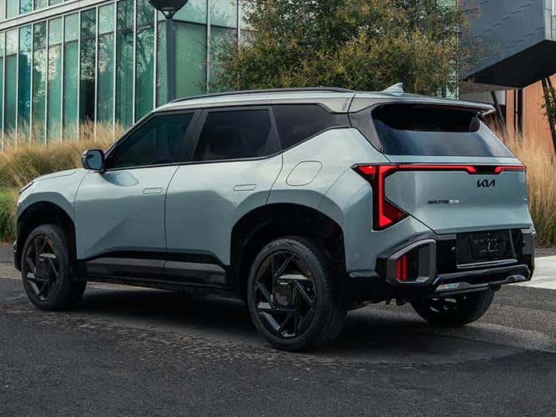A sleek, modern SUV with a light gray body is parked on a street. It features bold, angular tail lights and tinted windows, positioned near urban greenery.