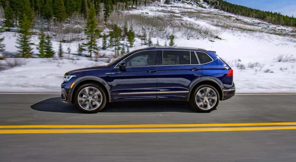 Side view of a blue 2024 Volkswagen Tiguan SEL R-Line driving past a snowy hillside.