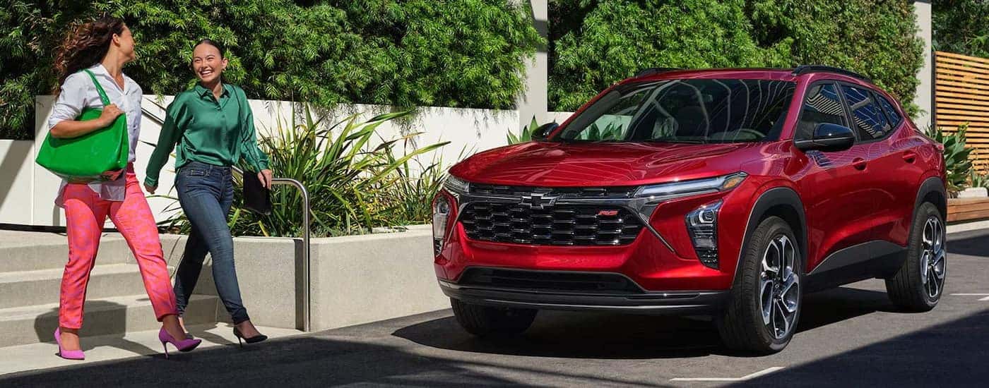 Two people walking beside a red 2024 Chevy Trax RS parked beside a curb.