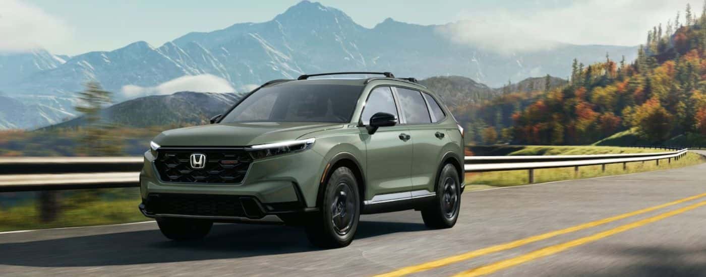 A green 2026 Honda CR-V TrailSport shown driving by a guard rail and mountains.