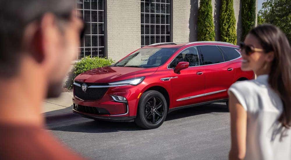 Two people are walking towards a red 2024 Buick Enclave parked on the side of a street.