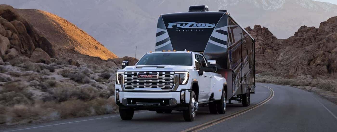 A white 2024 GMC Sierra 2500 HD Denali is shown towing a trailer on a highway.