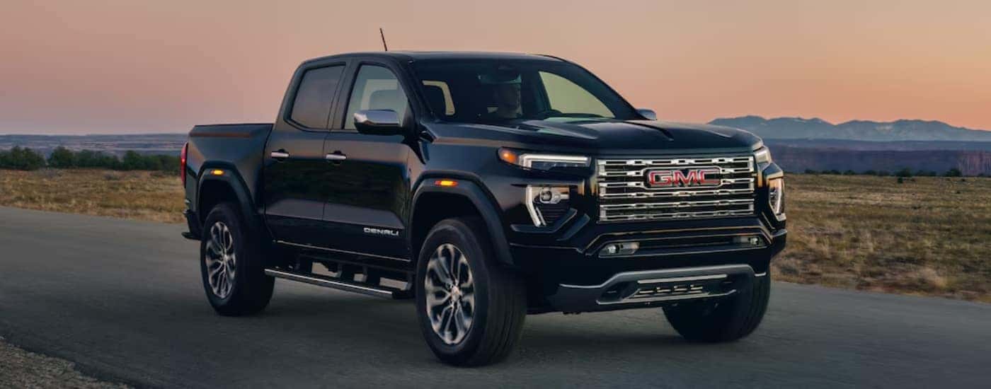 A black 2024 GMC Canyon Denali driving on an open road at sunset.