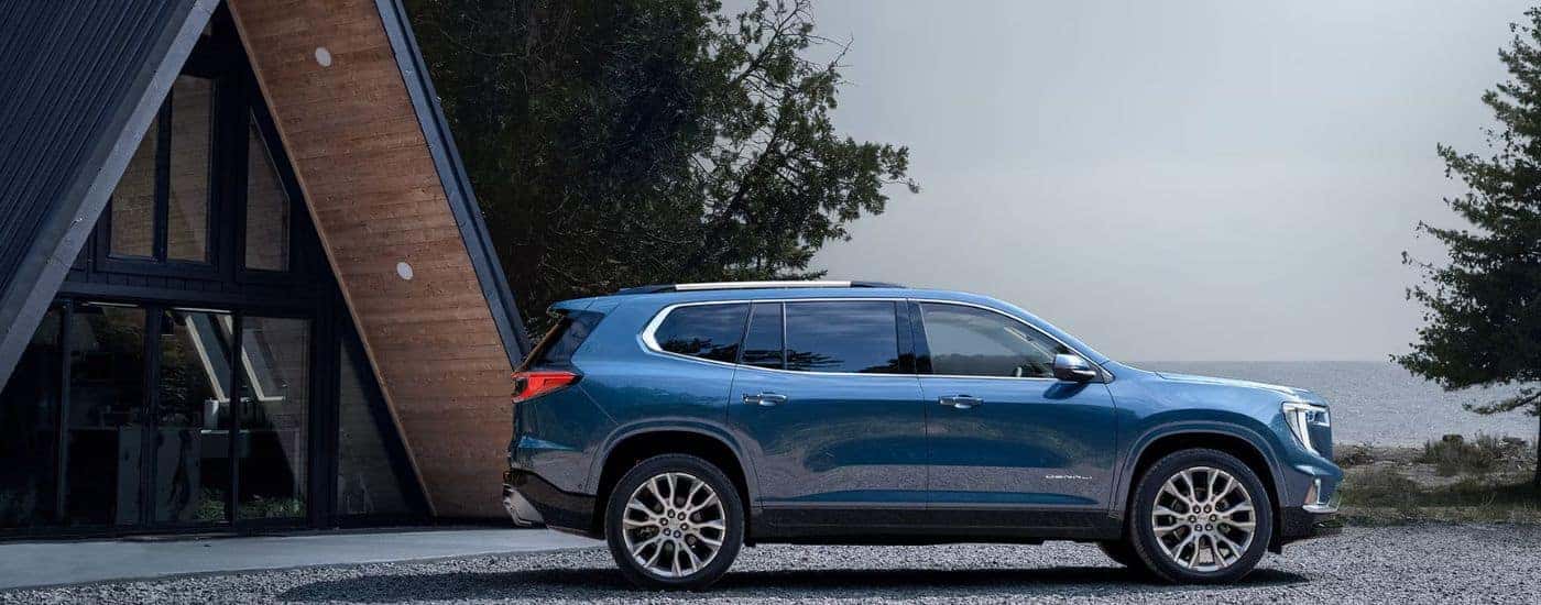 Blue 2026 GMC Acadia Denali parked in front of a lakeside cabin.