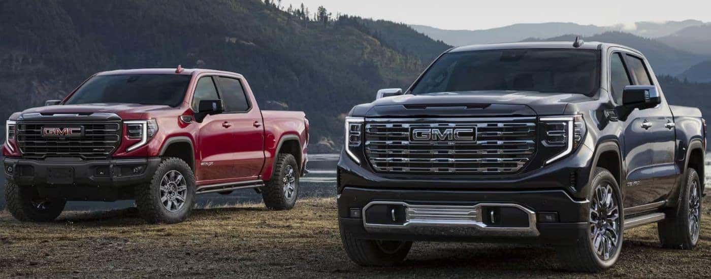A red and a black 2026 GMC Sierra 1500 parked in front of mountains and lake.