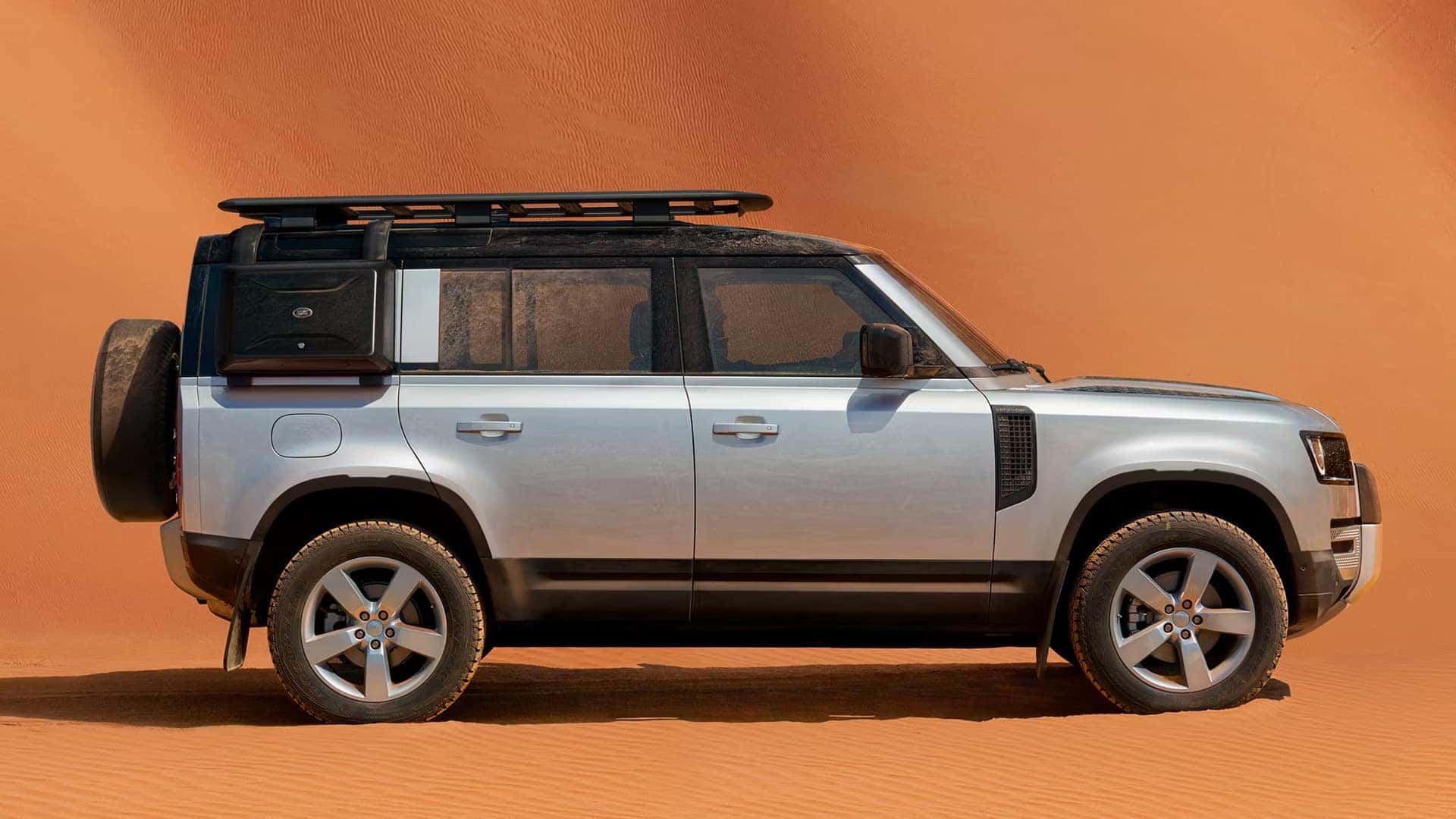 White Land Rover Defender with black roof parked on desert sand against orange/rust colored background