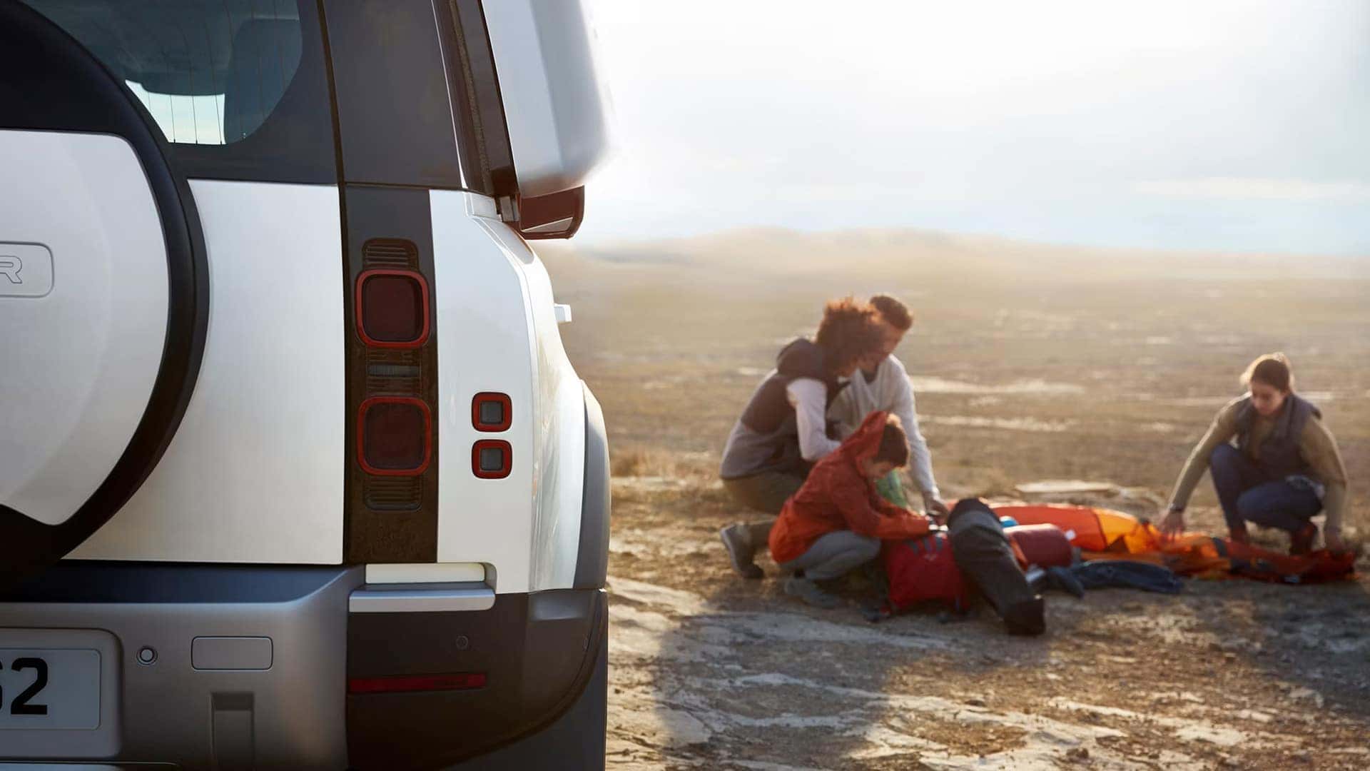 White Land Rover Defender parked in desert with people setting up camping equipment in the background