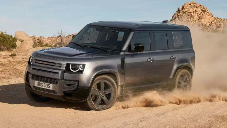 Silver Land Rover Defender driving through desert terrain with dust clouds around tires
