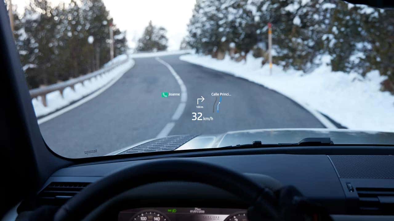 View from inside a vehicle showing an augmented reality navigation display on the windshield while driving on a snowy mountain road.