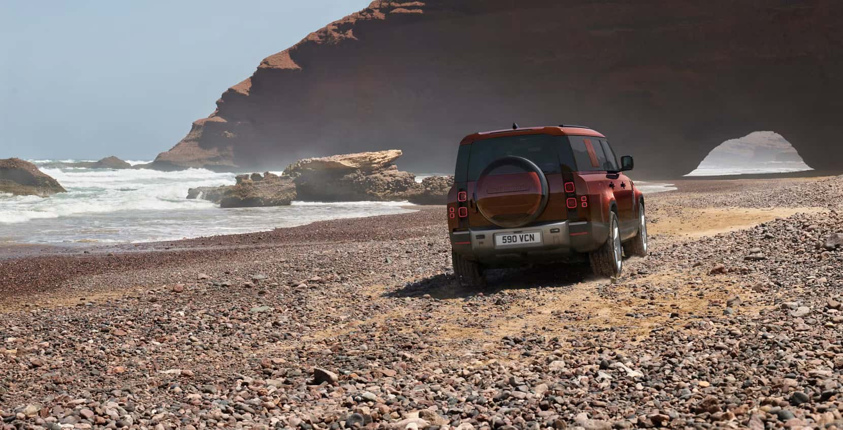 a reddish copper colored defender 130 driving on a rocky beach next to a rugged terrain and water