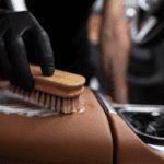 A person wearing black gloves scrubs a tan leather car seat with a brush and cleaning foam during an interior detailing.