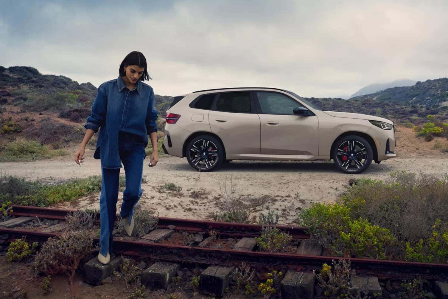 a lady walking over railroad tracks next to a bmw x3 parked on sandy gravel with a desert in the background
