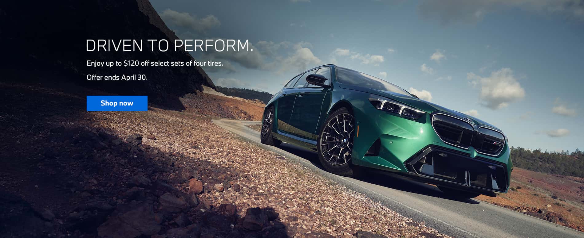 Front three-quarter view of a BMW M5 driving along a curved road, seen from a low, ground-level perspective. The camera looks upward toward the vehicle against a bright blue sky.