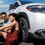Couple sitting on the ground beside a white 2025 Toyota Corolla Cross outdoors
