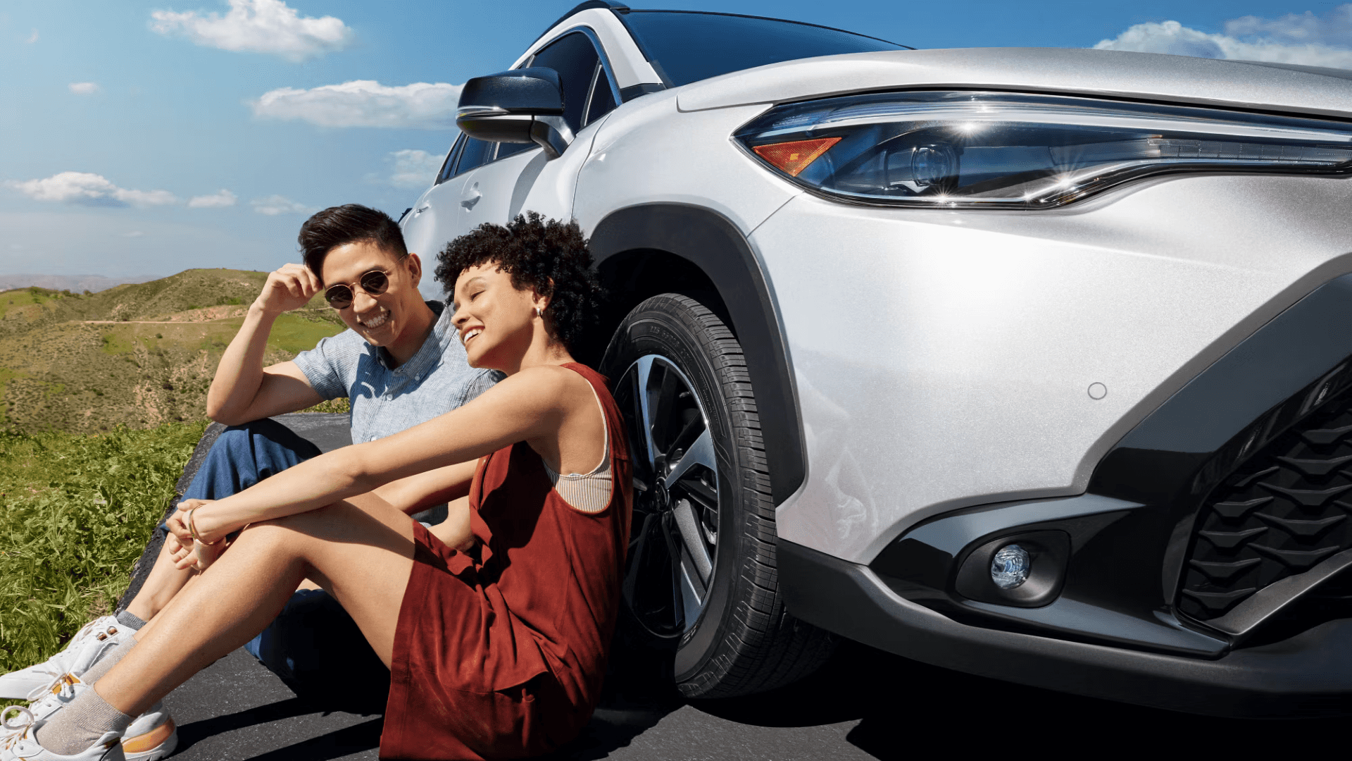 Couple sitting on the ground beside a white 2025 Toyota Corolla Cross outdoors